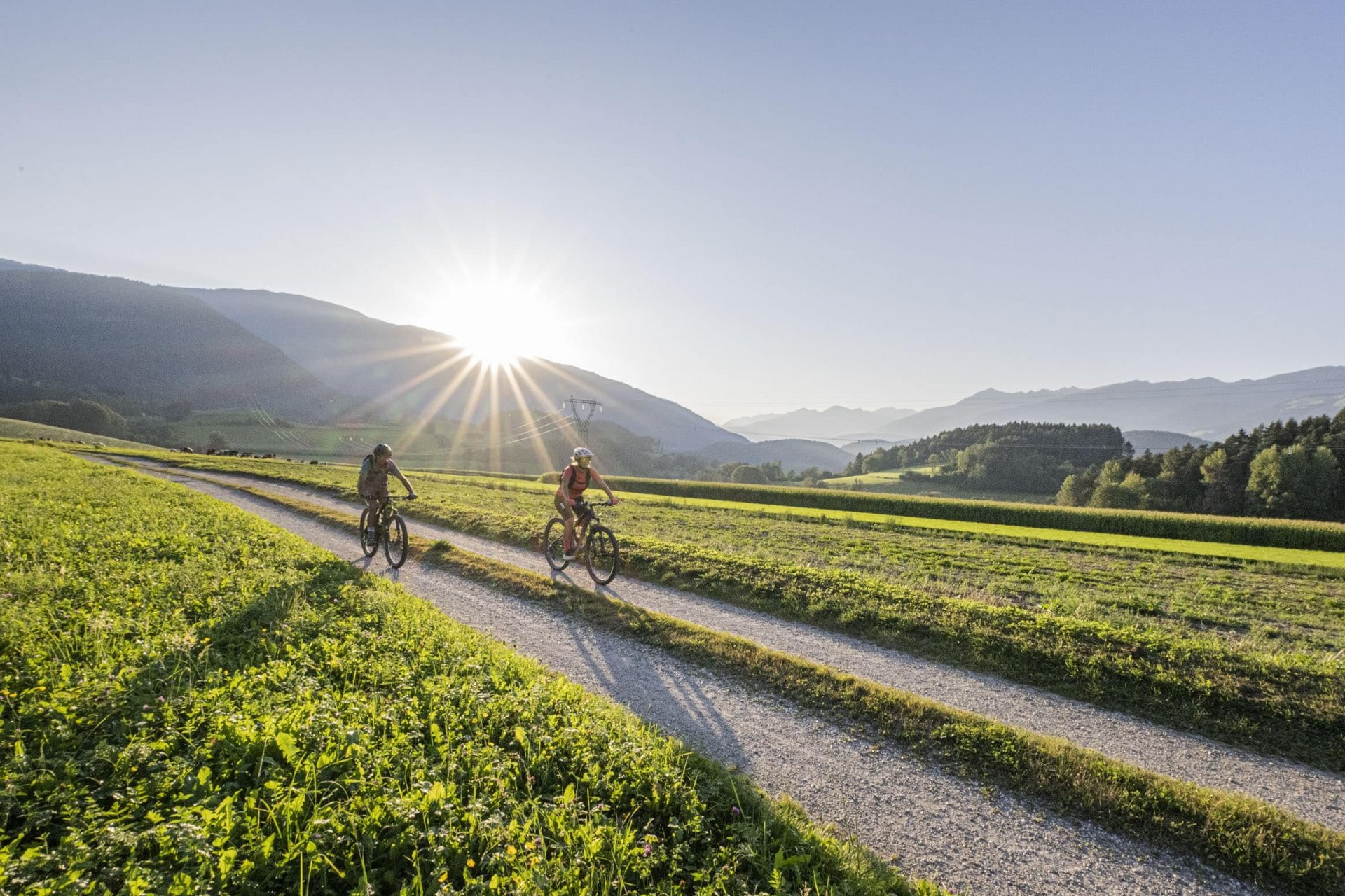 Velontour © Harald Wisthaler Zwei Radfahrer fahren auf einem Feldweg mit Bergen und Sonnenuntergang im Hintergrund