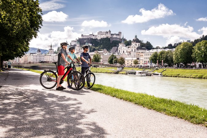 Velontour © Velontour Drei Radfahrer mit Helm an Flussufer vor Schloss und Stadt