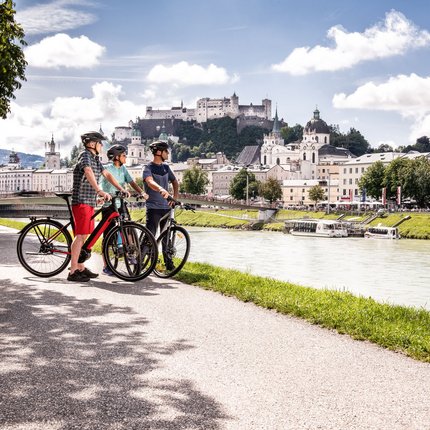 Drei Radfahrer mit Helm an Flussufer vor Schloss und Stadt
