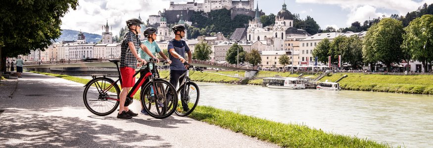 Radurlaub im Salzburger Land © Velontour Drei Radfahrer mit Helm an Flussufer vor Schloss und Stadt