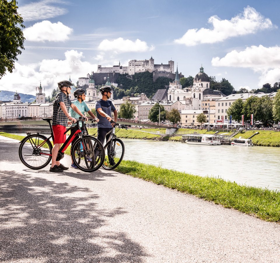Testblog © Velontour Drei Radfahrer mit Helm an Flussufer vor Schloss und Stadt