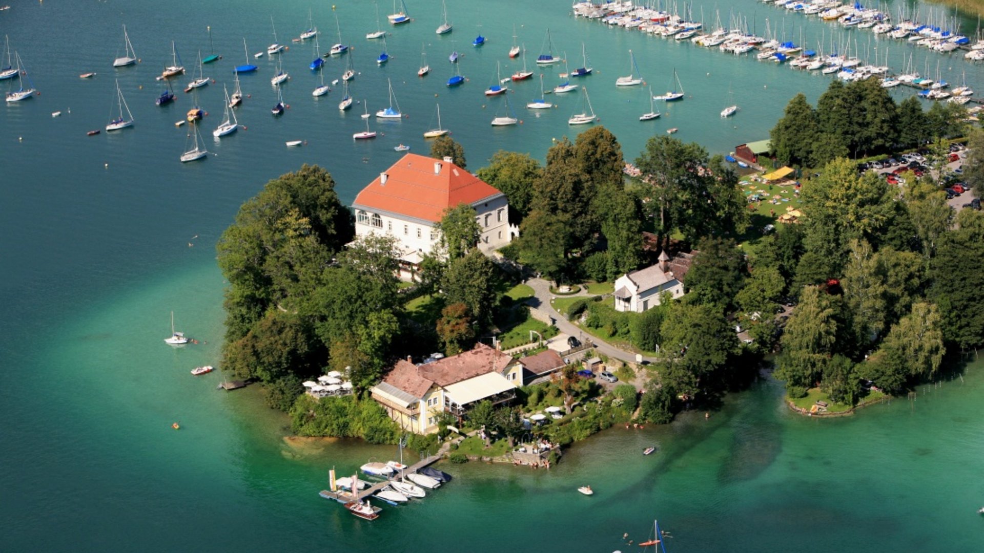 Schlössertour rund um Klagenfurt am Wörthersee Luftaufnahme einer bewaldeten Insel mit Gebäuden und Segelbooten im klaren Wasser